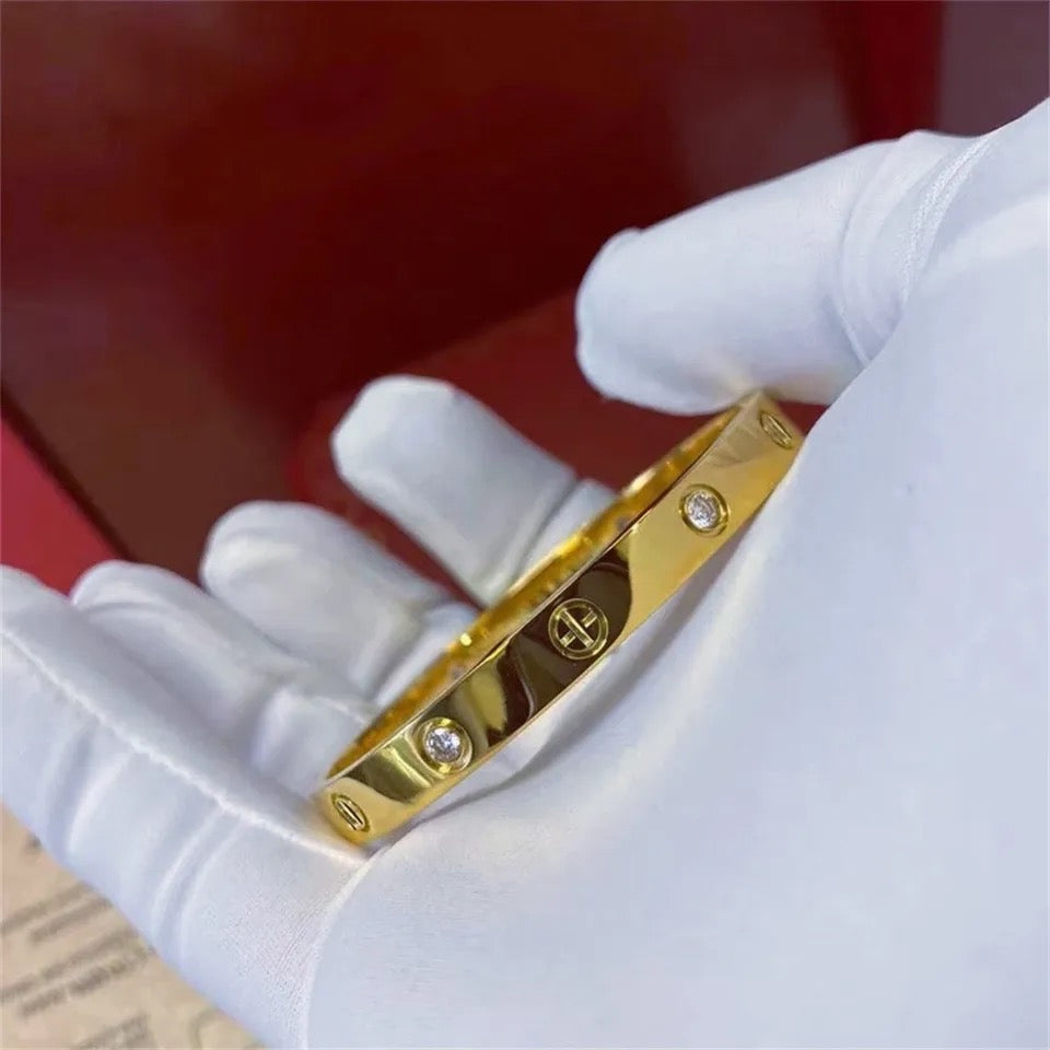 Close-up of gold bracelet with screw detailing and crystal accents, held in a gloved hand against red background.
