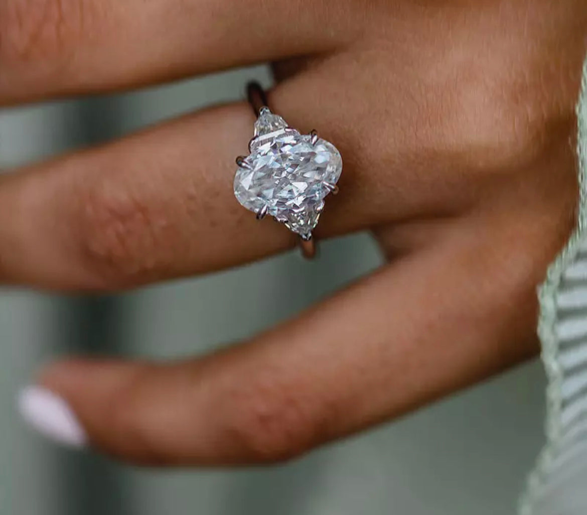 A hand holding a white oval-shaped Moissanite ring with a silver band and a yellow gold plating.