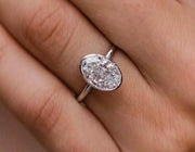 A hand displaying an oval-cut bezel-set moissanite ring on a silver 925 band with white gold plating.