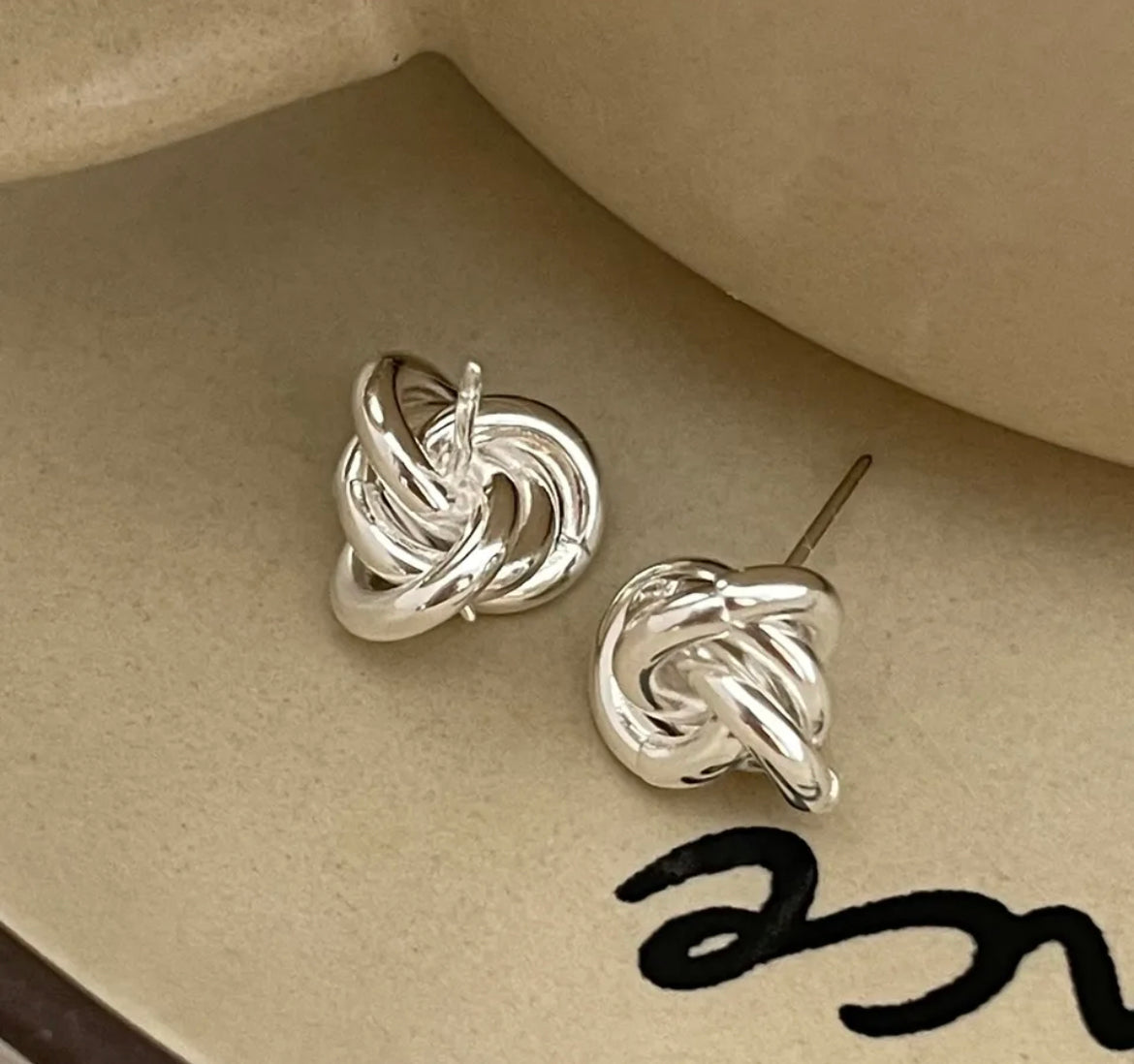 A pair of white gold plated knot earrings on a beige background with a close-up of the earring post showing the detail of the knot design.
