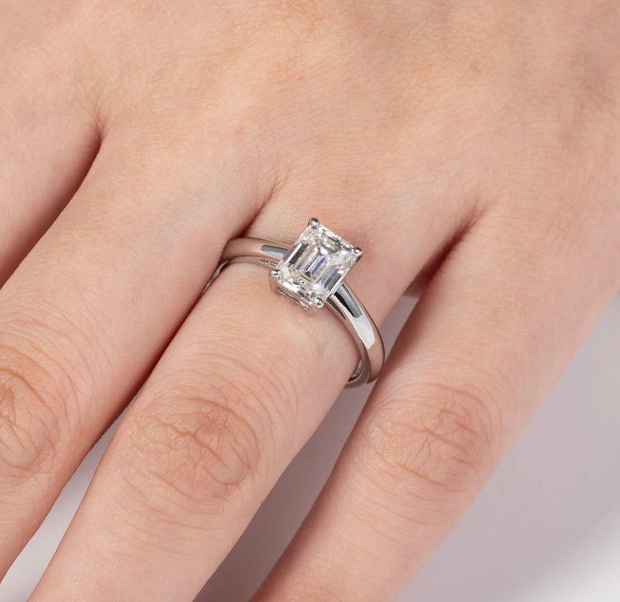 A hand displaying a silver-colored emerald cut moissanite ring on a finger.
