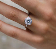A hand displaying a round bezel set Moissanite solitaire ring on a finger, with options for metallic colors including silver, yellow, and white gold mentioned in the product description.