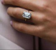 A hand holding a sparkling emerald cut bezel set moissanite ring with a gold band, indicating it is a engagement or wedding ring.
