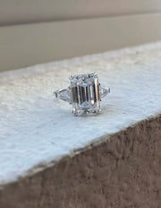 An 8ct emerald cut Moissanite ring with tapered baguettes, displayed on a white surface, with options for silver and gold plating.