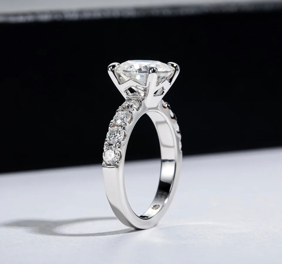 A silver ring with a central moissanite stone and smaller stones set in a pave around the band, displayed against a black background.