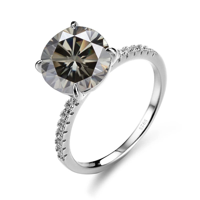 A silver ring featuring a central Moissanite diamond, surrounded by smaller clear stones set in a classic four-clawed setting.