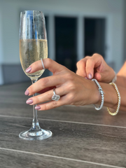 Two Soho 4mm Moissanite Tennis Bracelets in gold and silver worn together on wrist, paired with a clover ring and champagne glass