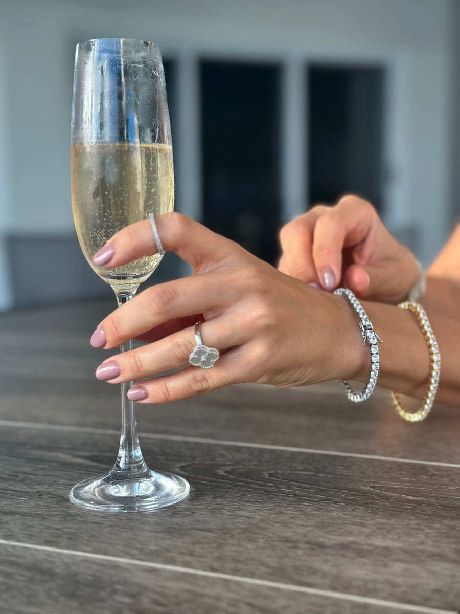 Two Soho 4mm Moissanite Tennis Bracelets in gold and silver worn together on wrist, paired with a clover ring and champagne glass