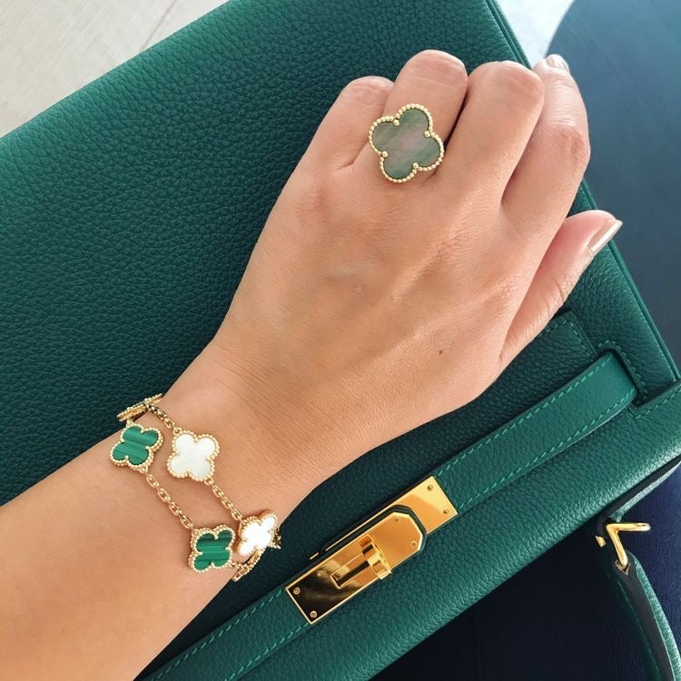 A person's wrist wearing a green clover bracelet with gold-colored beads and a gold-colored clasp, next to a green purse.