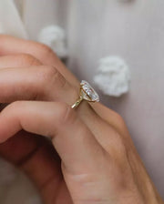 A hand holding a 3ct crushed ice hybrid cut Moissanite ring, with a silver band, over a blurred background of a white fabric.