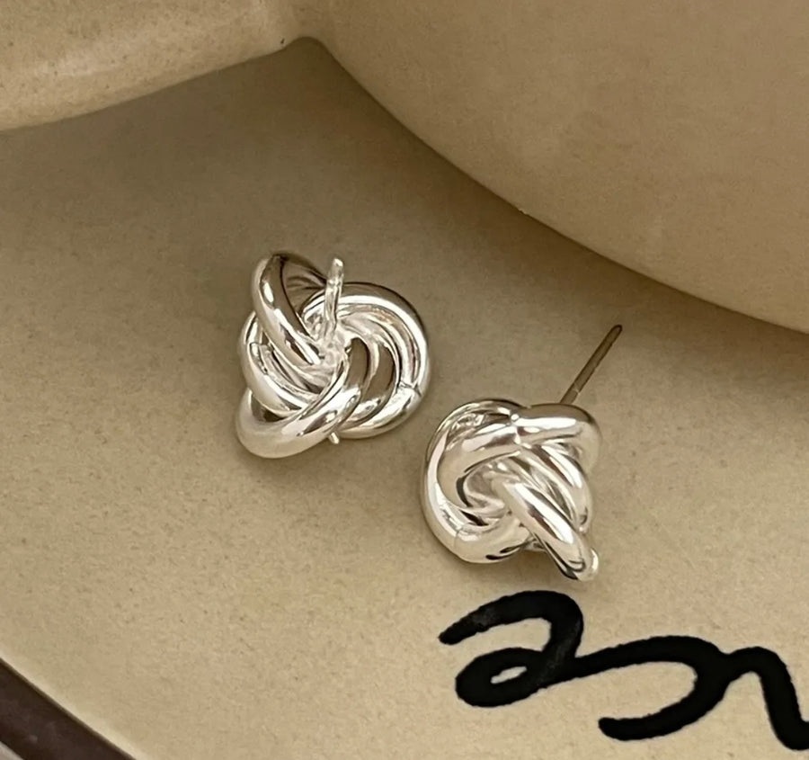 A pair of white gold plated knot earrings on a beige background with a close-up of the earring post showing the detail of the knot design.