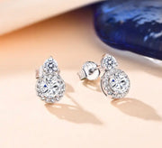 A pair of white Moissanite earrings set in silver, with a total weight of 1ct each, displayed against a blue textured background.