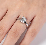 A hand displaying a silver-colored emerald cut moissanite ring on a finger.