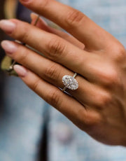 A close-up image of a person's hand showcasing a silver ring with a large central crushed ice hybrid Moissanite gemstone, surrounded by a smaller halo of stones.