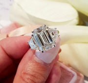 A hand holding a 5ct emerald cut baguette moissanite ring with a silver band and a plated white gold shank.